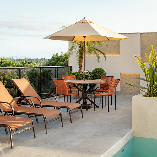 Na imagem é possível ver a piscina do residencial Ilha do Mel construído pela Itapuí Incorporadora. No local, temos um ambiente com mesa rodeada por 4 cadeiras em tons terrosos. Há também 3 cadeiras de piscina na cor terracota próximas a beira da piscina.