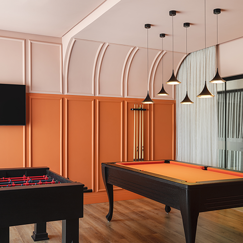 na imagem é possível ver a sala de jogos do residencial ilha do mel. as paredes são pintadas nas cores terracota e branco, divididas numa proporção de 70/30, e com acabamentos com boiserie. Há duas mesas de sinuca, com pendentes em cima criando um clima intimista pra imagem.