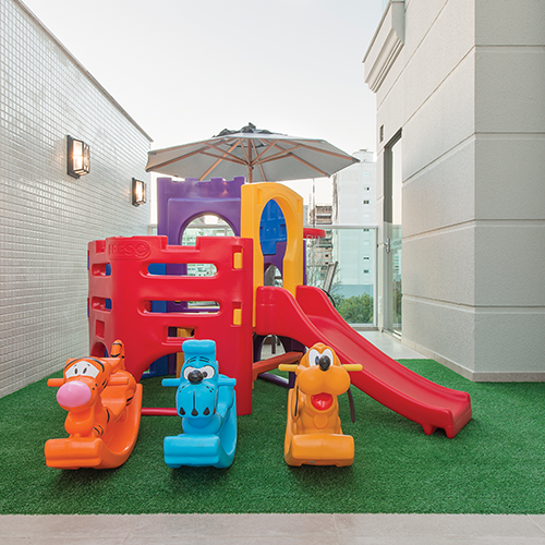 playground do residencial Lago Negro construído pela Itapuí Incorporadora. Há na imagem um escorregador com escadinhas, cadeirinhas de balanço e um guarda-sol em cima de um local gramado.