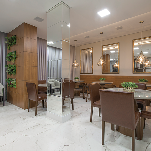 na imagem é possível ver o salão de festas do residencial Lago Negro, construído pela itapuí incorporadora. A imagem apresenta o ambiente com piso que simula mármore branco, revestimentos em madeira nos pilares e paredes pintadas em tons claros. Há mesas e cadeiras em formato redondo espalhadas pelo ambiente na cor marrom