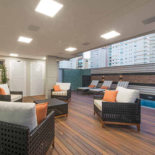 Área externa de lazer do residencial Lago Negro construído pela Itapuí Incorporadora. No local há piso simulando madeira escura, poltronas trançadas marrom escuro com estofado claro e almofadas laranjadas. Ao fundo é possível ver uma piscina e cadeiras de piscina beges com almofadas marrom