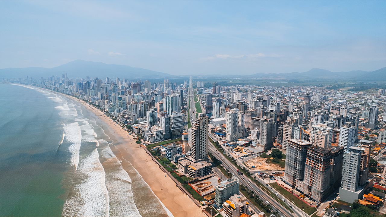 Vista aérea da orla de Itapema, com prédios à beira-mar e ondas quebrando na praia.