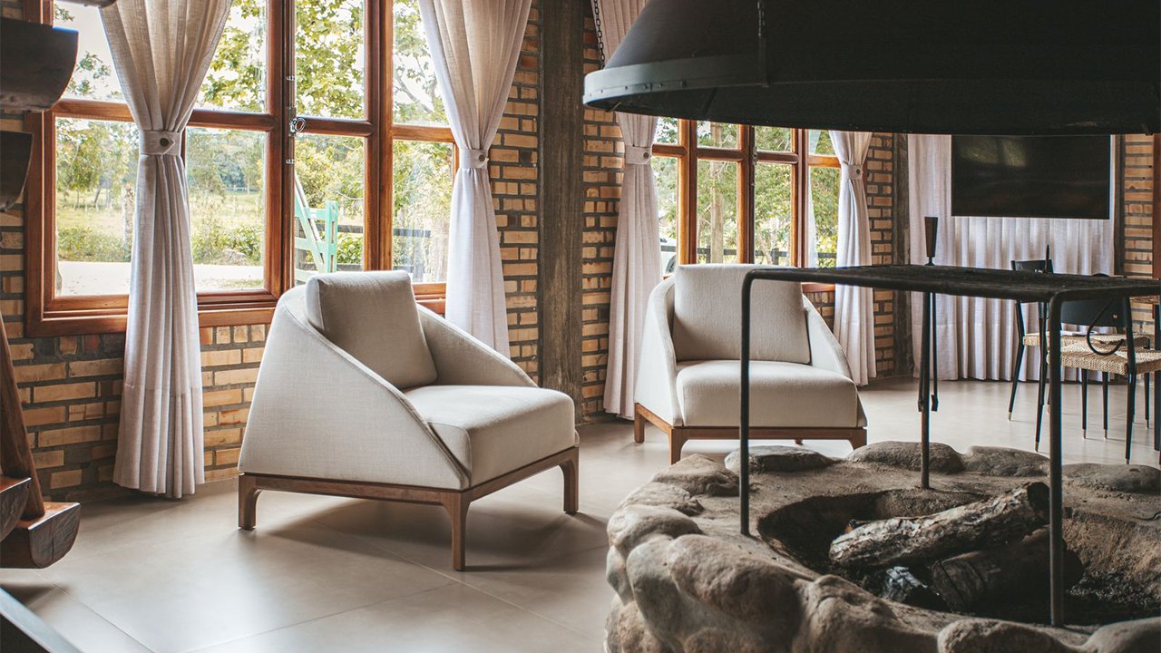 Sala com poltronas de linho claro, lareira rústica de pedra e iluminação natural – exemplo de tendências de decoração para o inverno com aconchego e materiais naturais.