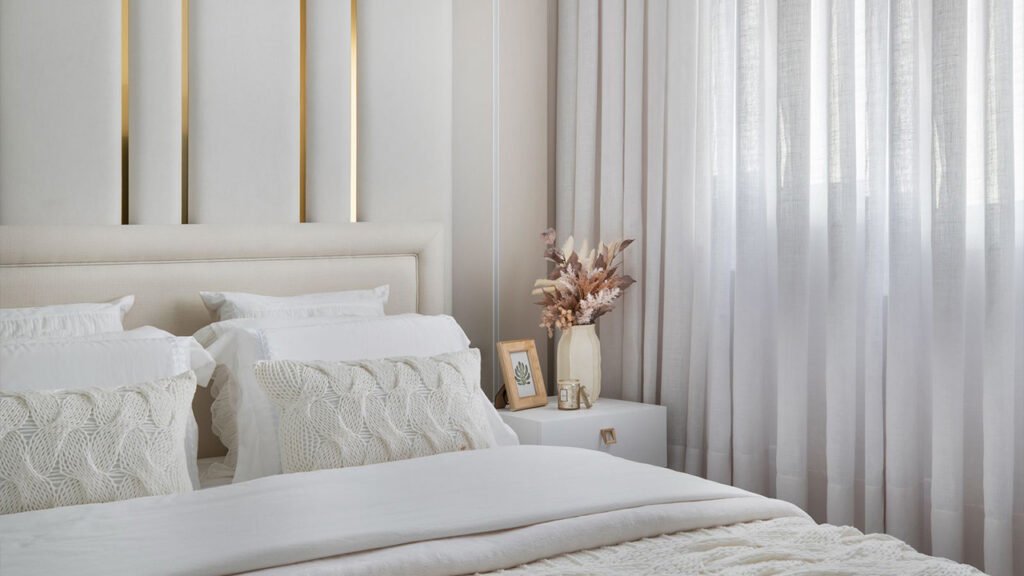 Quarto decorado com tons neutros, almofadas em tricô e cortinas leves, refletindo as tendências de decoração para o inverno 2025 com foco no toque e no conforto visual.